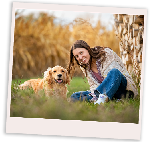 Erin - Support for Caregivers Ontario Erin and her dog Penny sit in a field, with corn in the back and wood stacked to the right. They face the camera and are smiling.